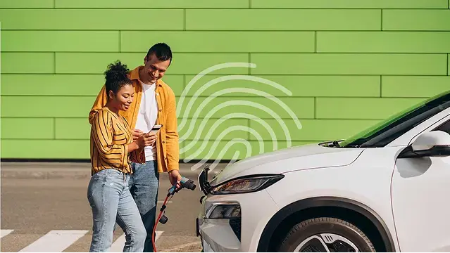 Een koppel bekijkt samen een smartphone naast een witte elektrische wagen die oplaadt voor een groene muur.