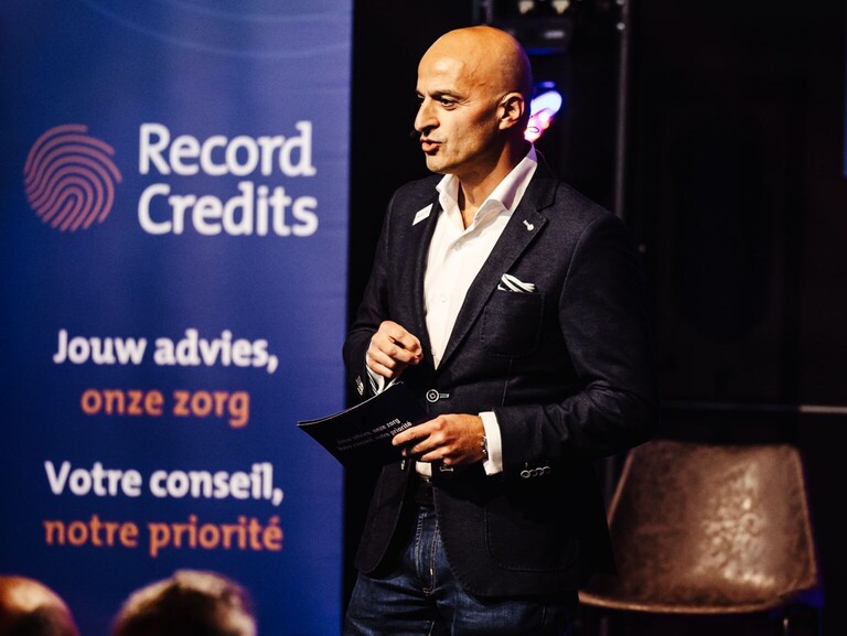 Turhan Özgüner spreekt op een podium voor een banner met de slogan Jouw advies, onze zorg.